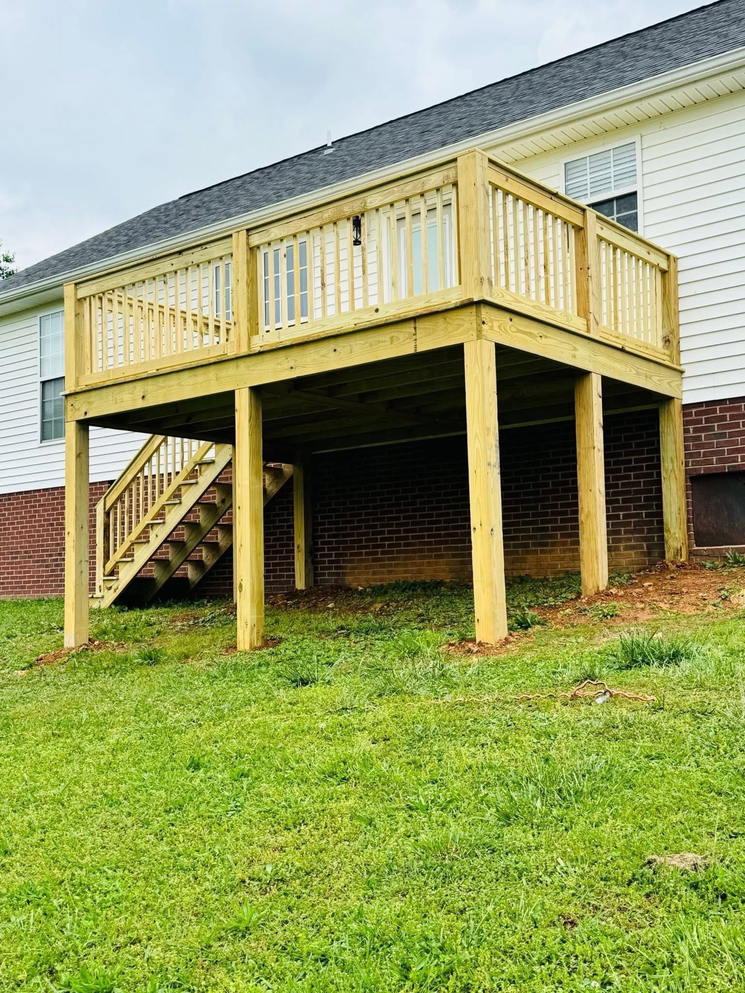 Raised wooden deck with stairs beside a house, supported by posts over a grassy yard.