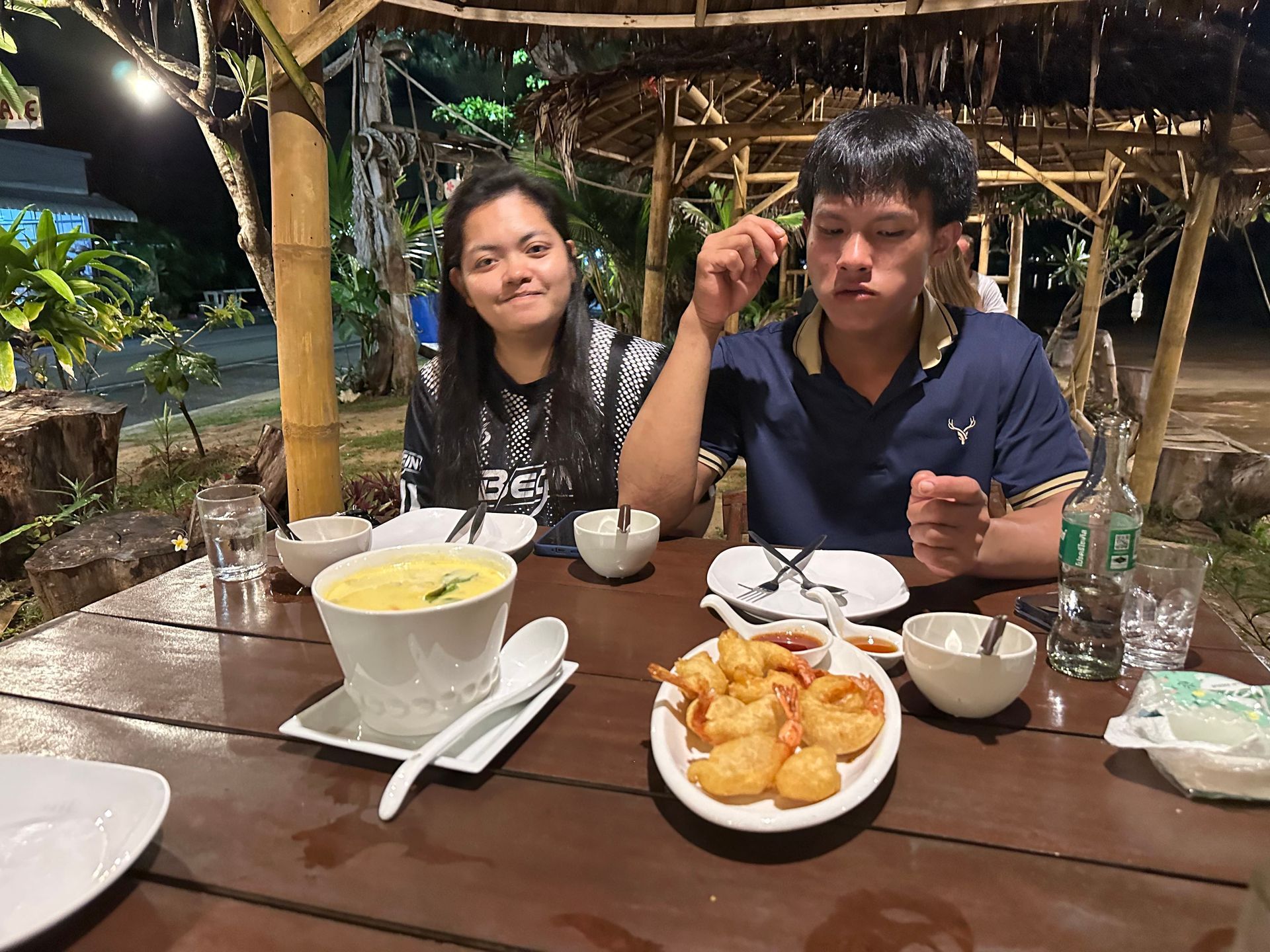 Two people at a wooden table outdoors with drinks and snacks under warm lights