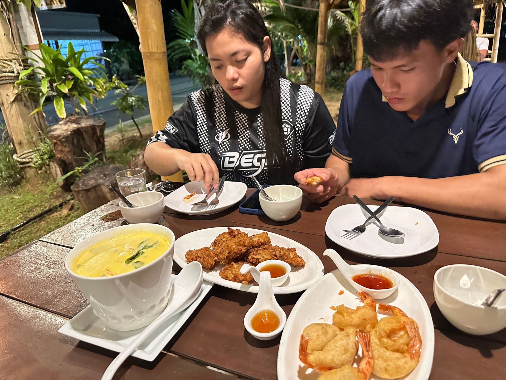 Two people eating a meal at a restaurant table with soup, fried dishes, and dipping sauces