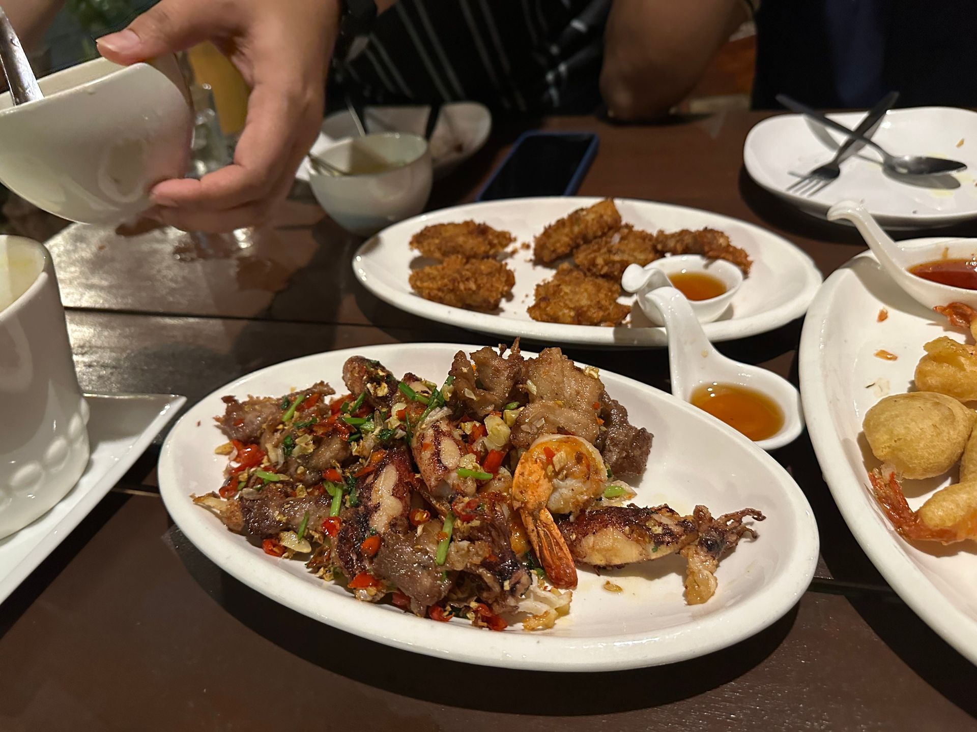 Hands serving Thai dishes on a table with bowls, sauces, and fried items