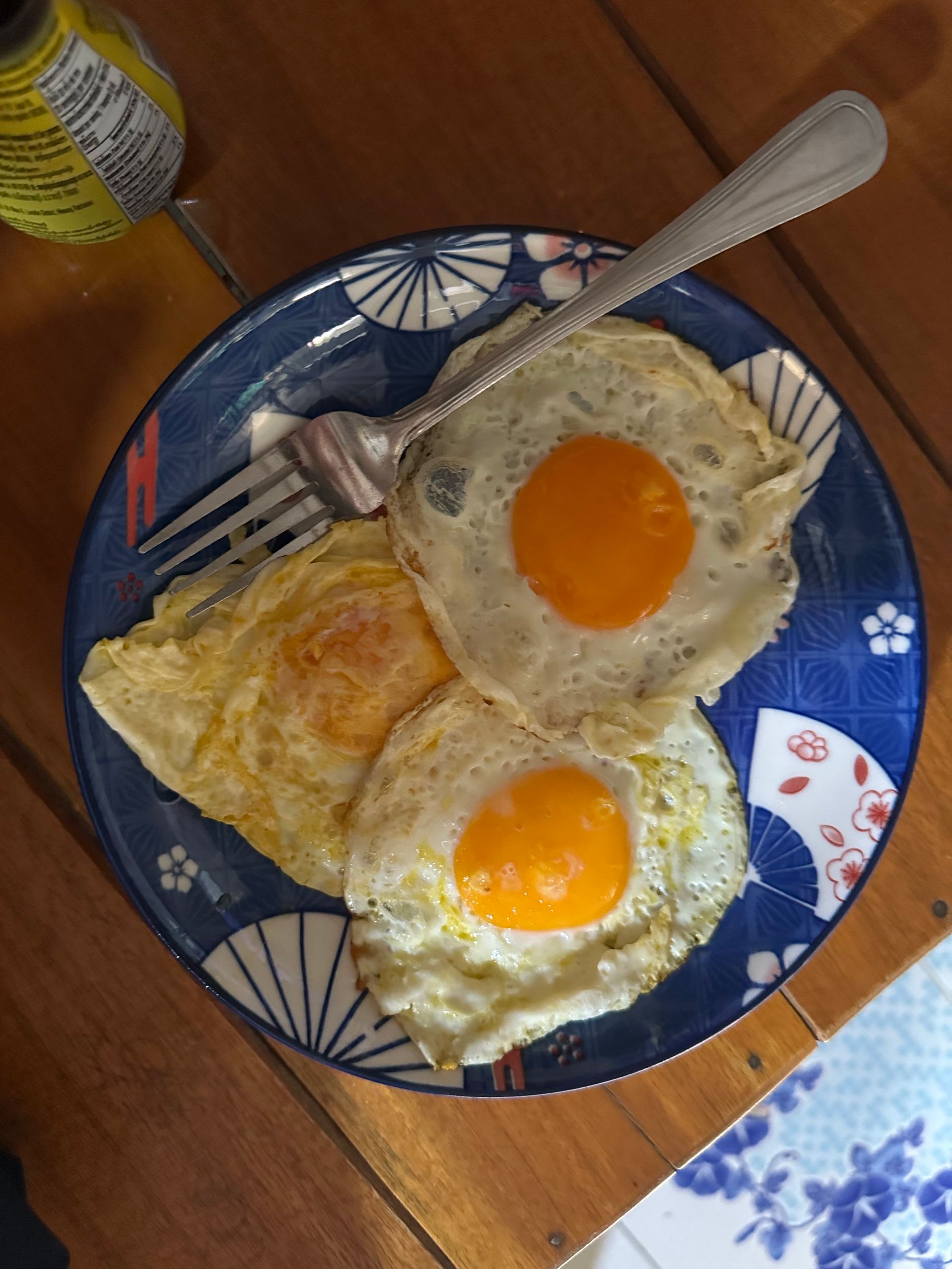 Plate with two fried eggs and a fork on a wooden table beside a drink bottle.