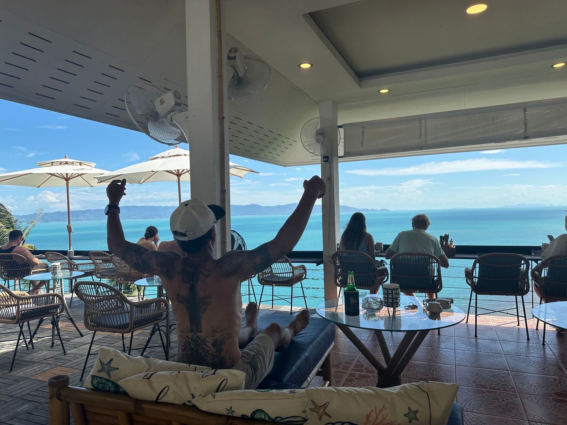 People sitting on a seaside patio under a covered roof, with one person stretching arms up toward the ocean.