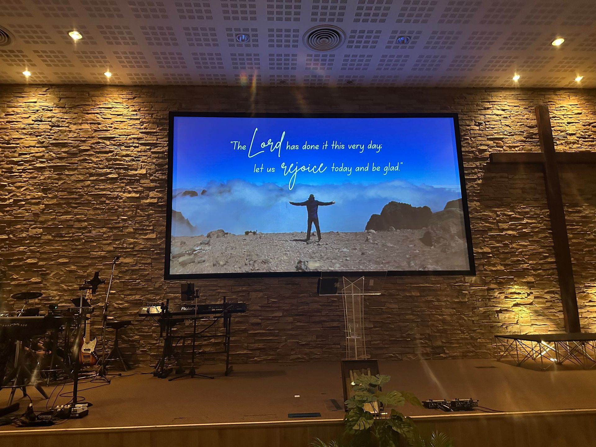 Indoor stage with stone wall backdrop and a large screen showing a person in a desert landscape under blue sky.
