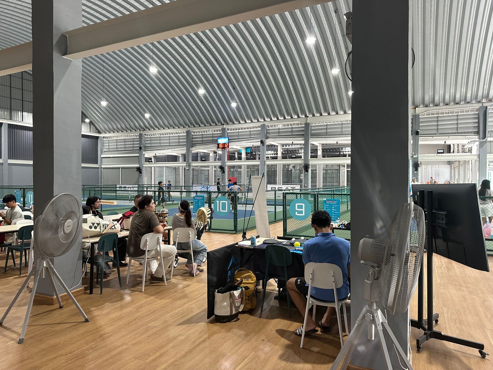 Indoor airport waiting area with seated travelers, ceiling lights, and large fans near glass walls.