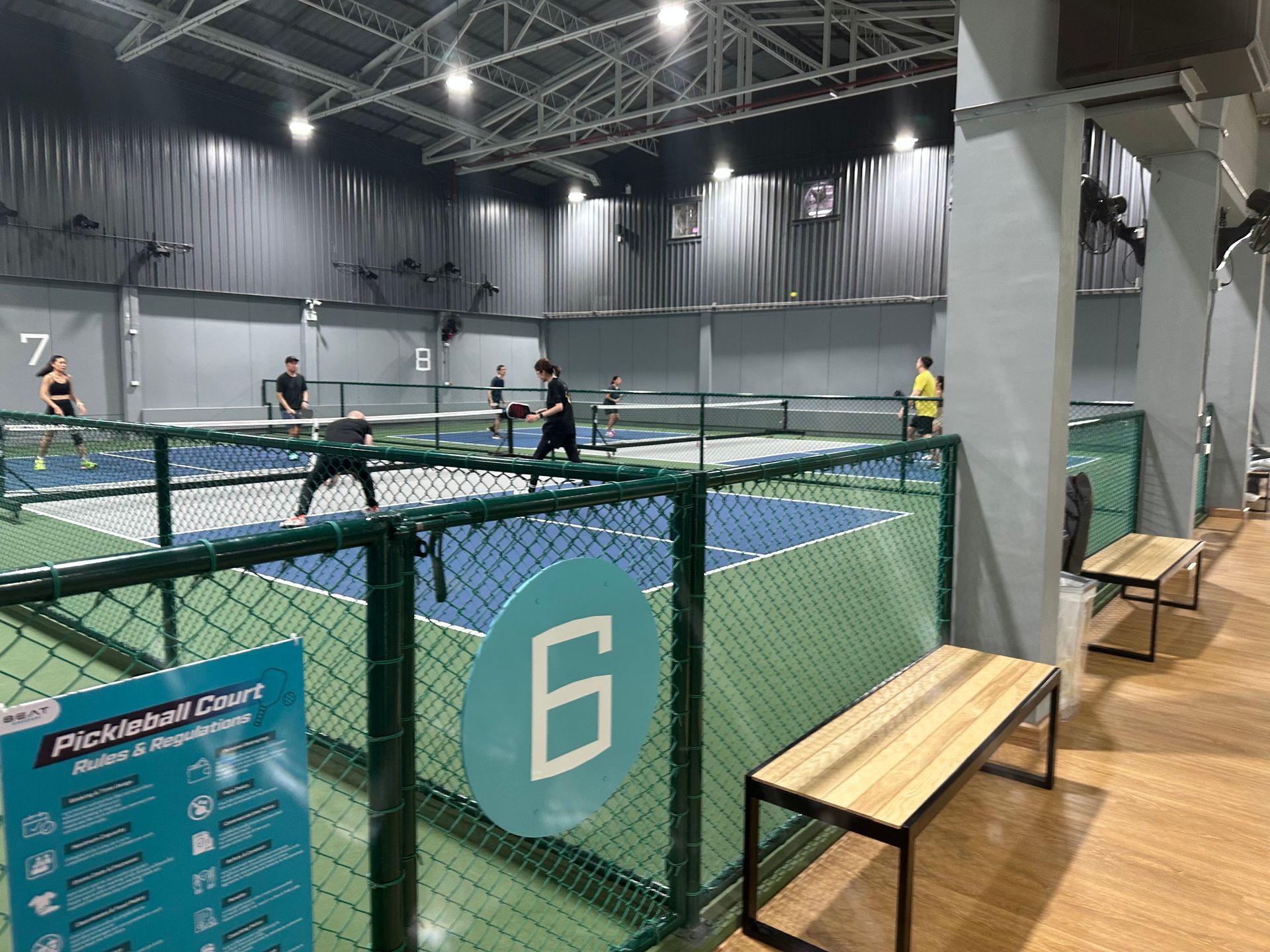 Indoor pickleball court with players, green fencing, and benches under bright overhead lights.