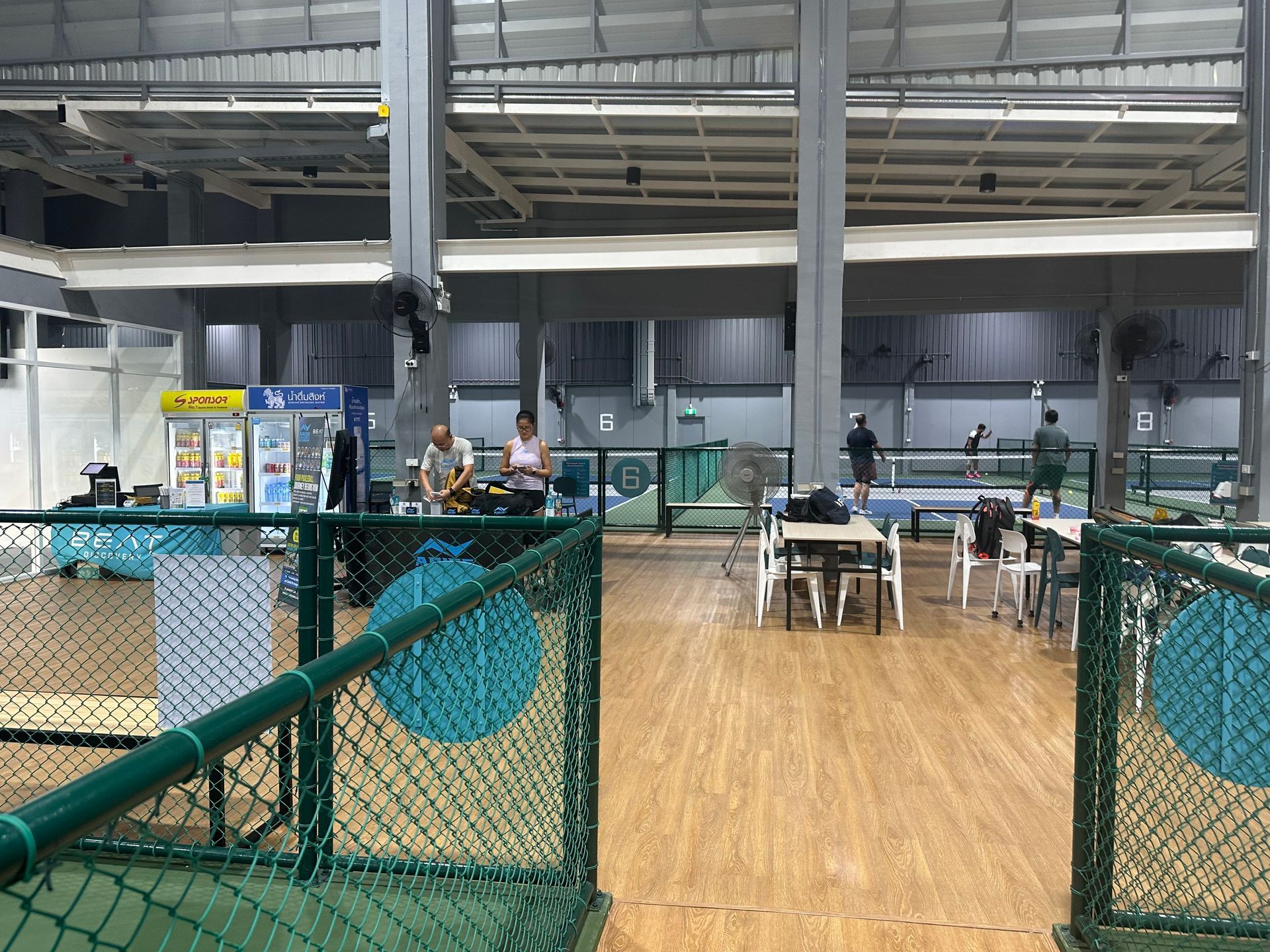 Indoor sports court with green fencing, wooden floor, and people seated at tables in the background