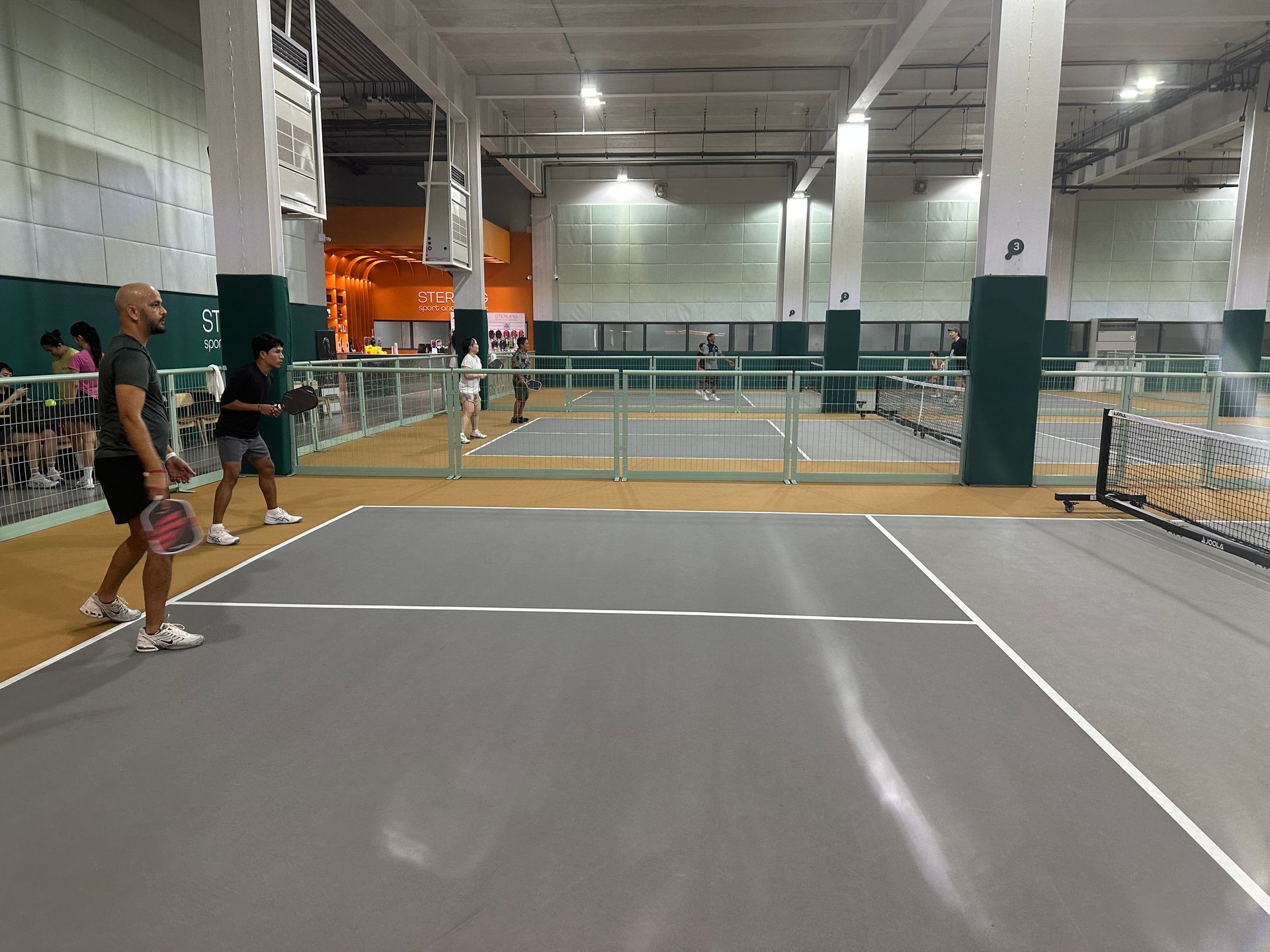 Indoor pickleball court with players at the net and one player in red shorts serving from the baseline