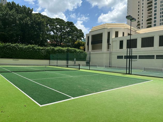 Outdoor court with green turf, fenced net area, and buildings in the background under a partly cloudy sky