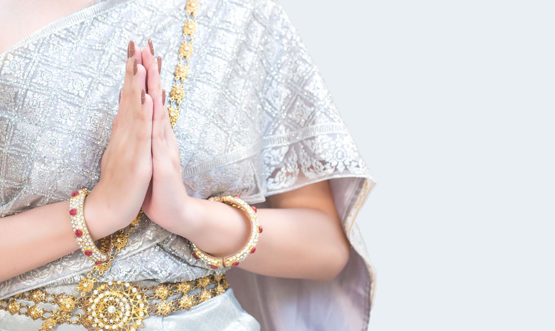 Person in white traditional attire with hands pressed together in a greeting gesture, wearing gold jewelry.