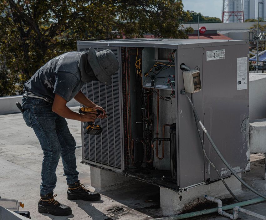 Technician repairing an outdoor HVAC unit on a rooftop, with tools and open panels exposed.