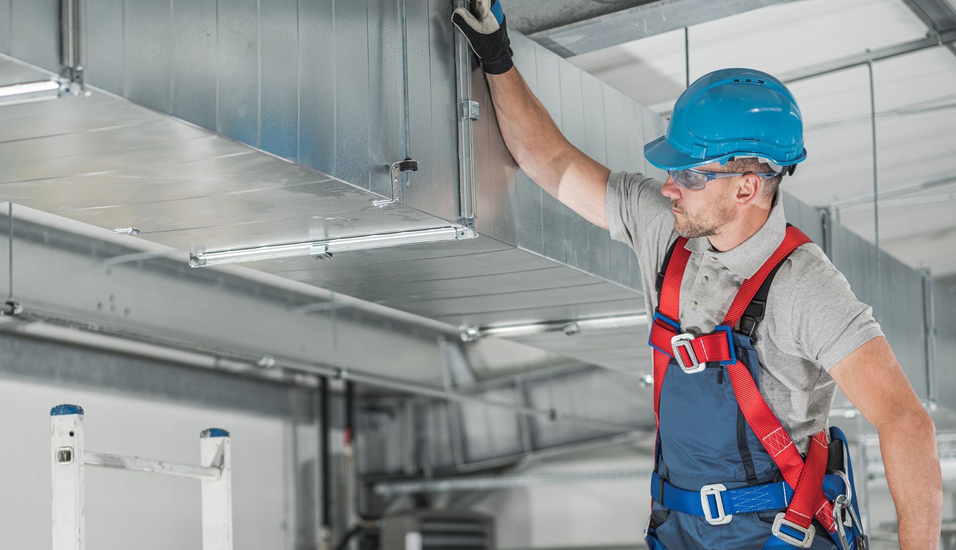 Worker in blue hard hat and safety harness inspecting overhead ductwork in an industrial facility