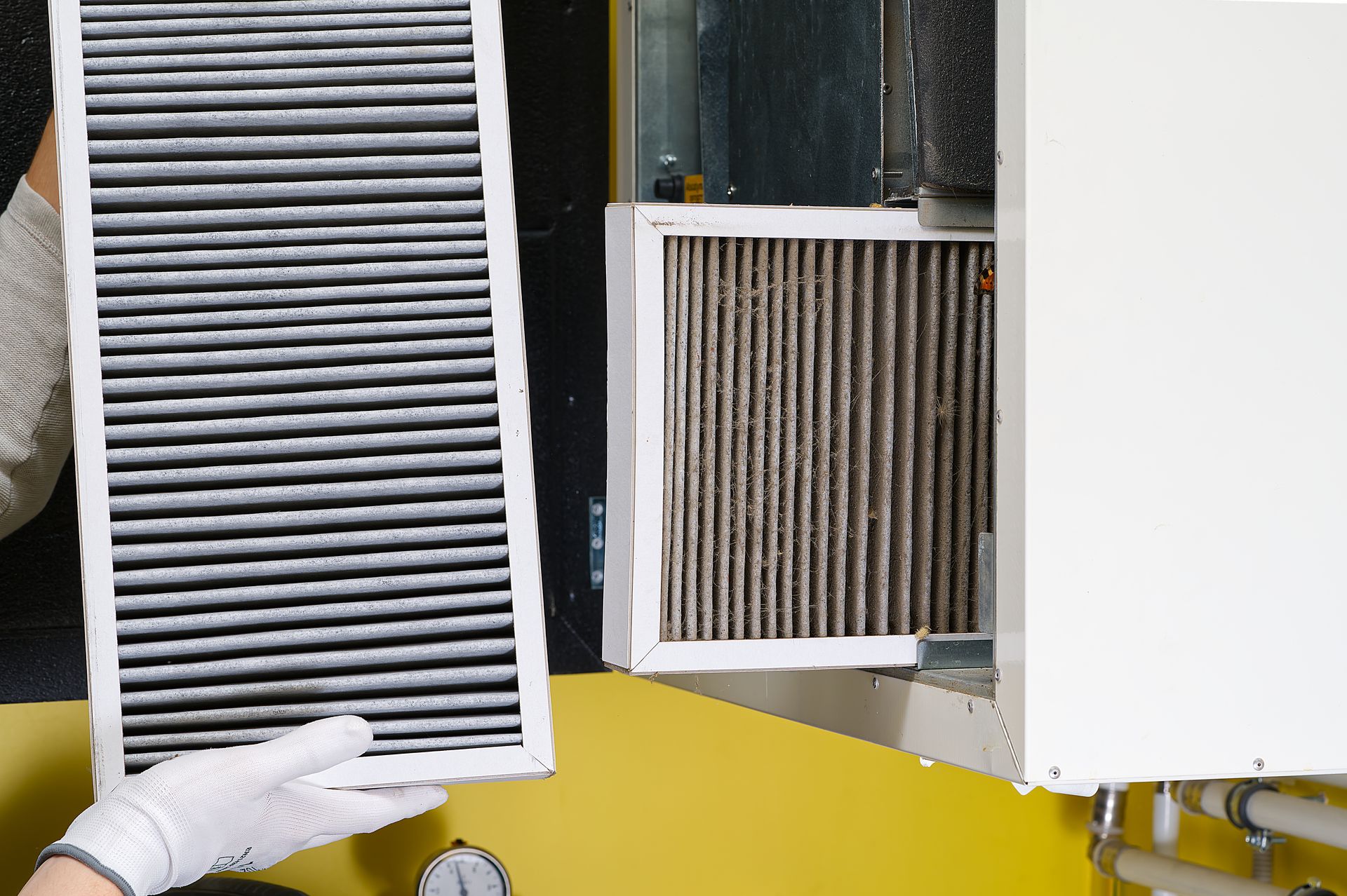 Gloved hand holding a white air filter beside an open HVAC unit with a dirty filter inside