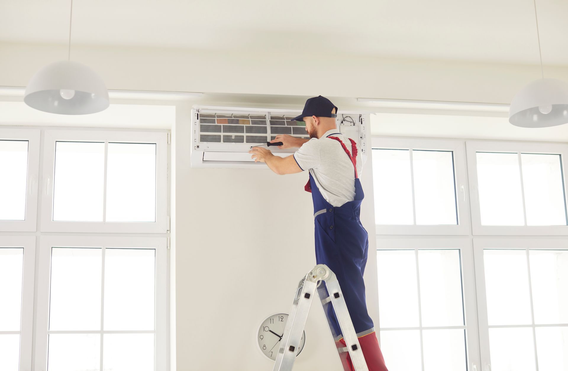 Technician standing on a ladder cleaning an air conditioner vent in a bright room