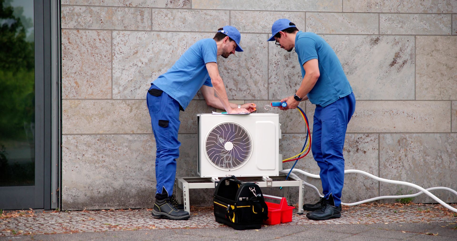 Two technicians in blue uniforms inspecting an outdoor air conditioning unit against a wall.