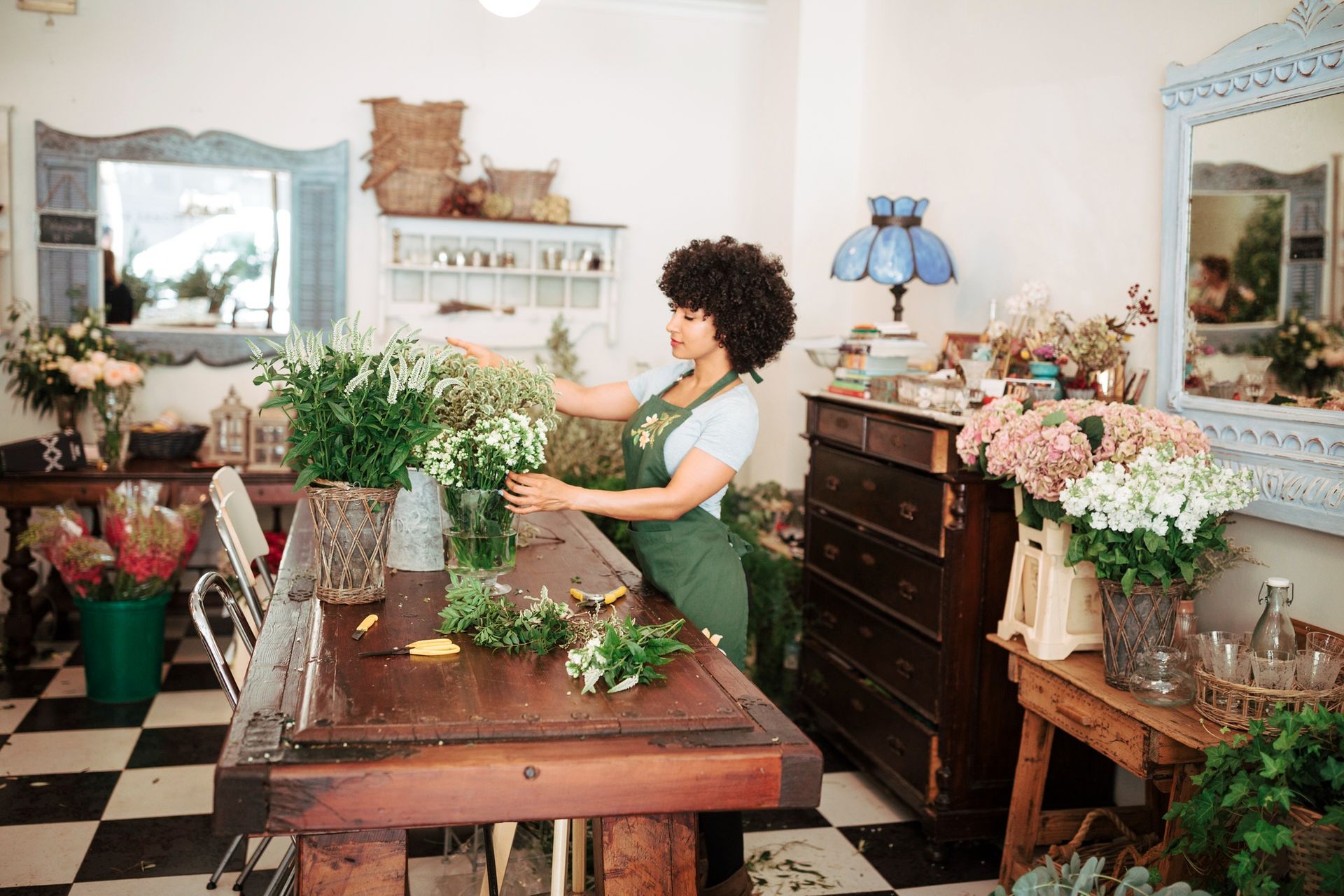 Woman arranging flowers at a wooden table in a flower shop. She wears a green apron and has curly hair.