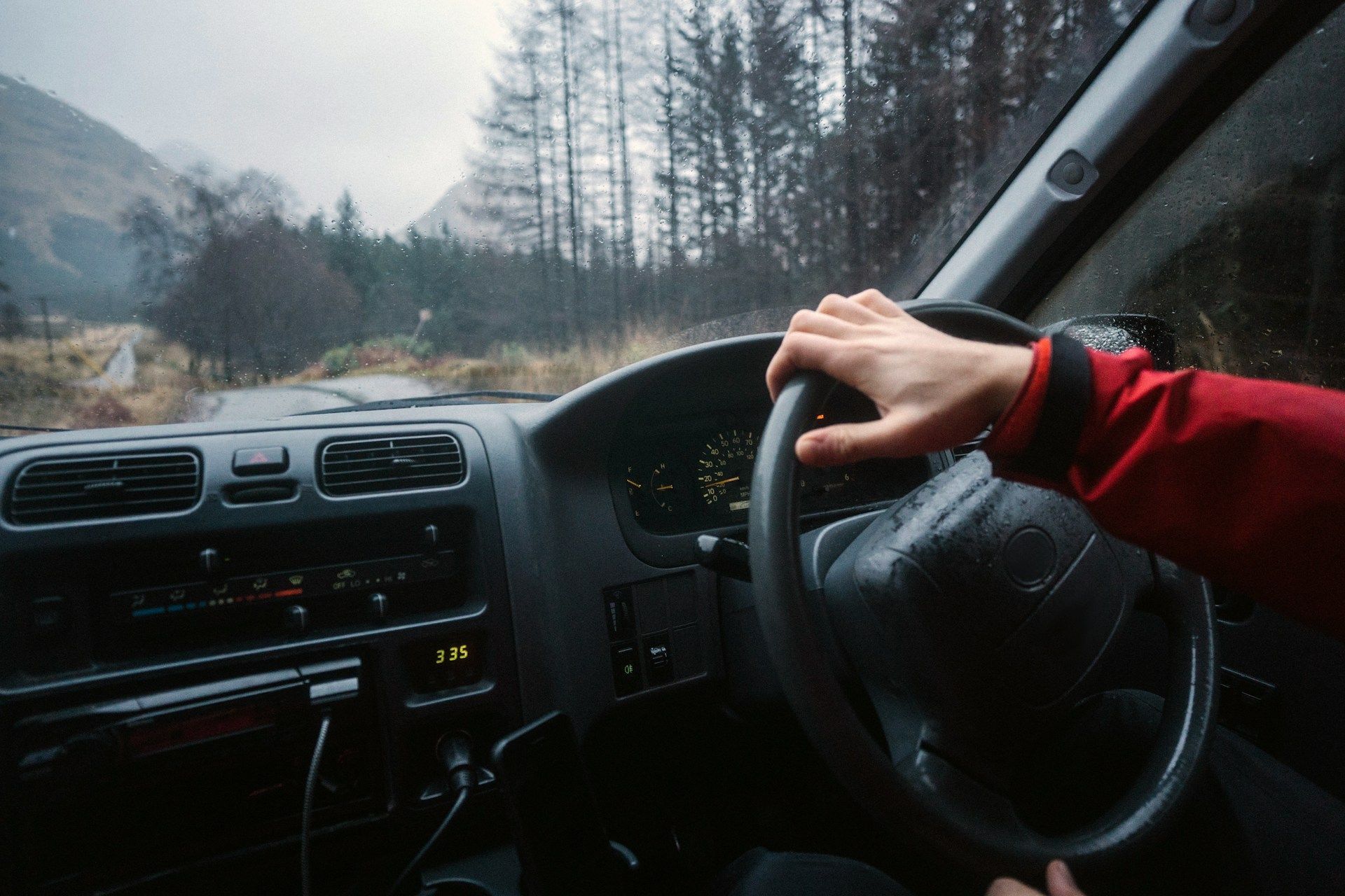 Hand gripping steering wheel inside a car, driving on a rainy road with a forest and mountains in the background.