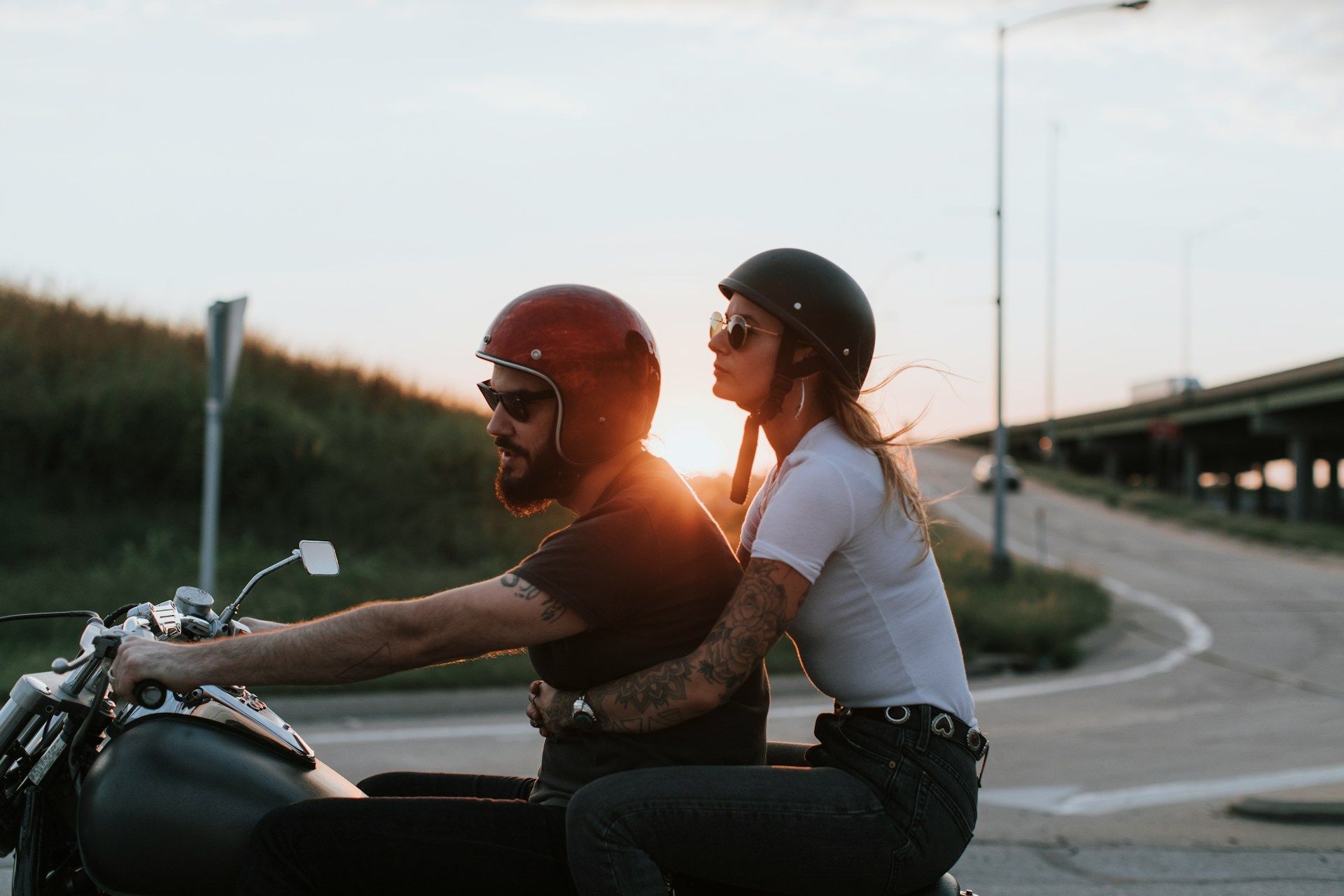 Man and woman on a motorcycle, wearing helmets and sunglasses, riding along a road at sunset.