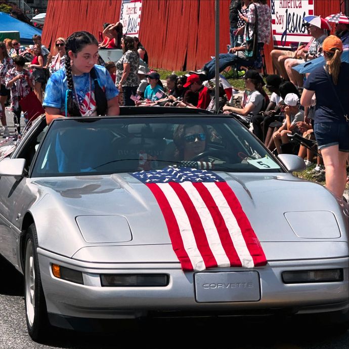 Silver convertible Corvette with red-white-blue stripes at a crowded outdoor event.