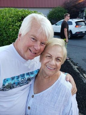 Two smiling people posing outdoors, one with an arm around the other, with a parked car in the background.