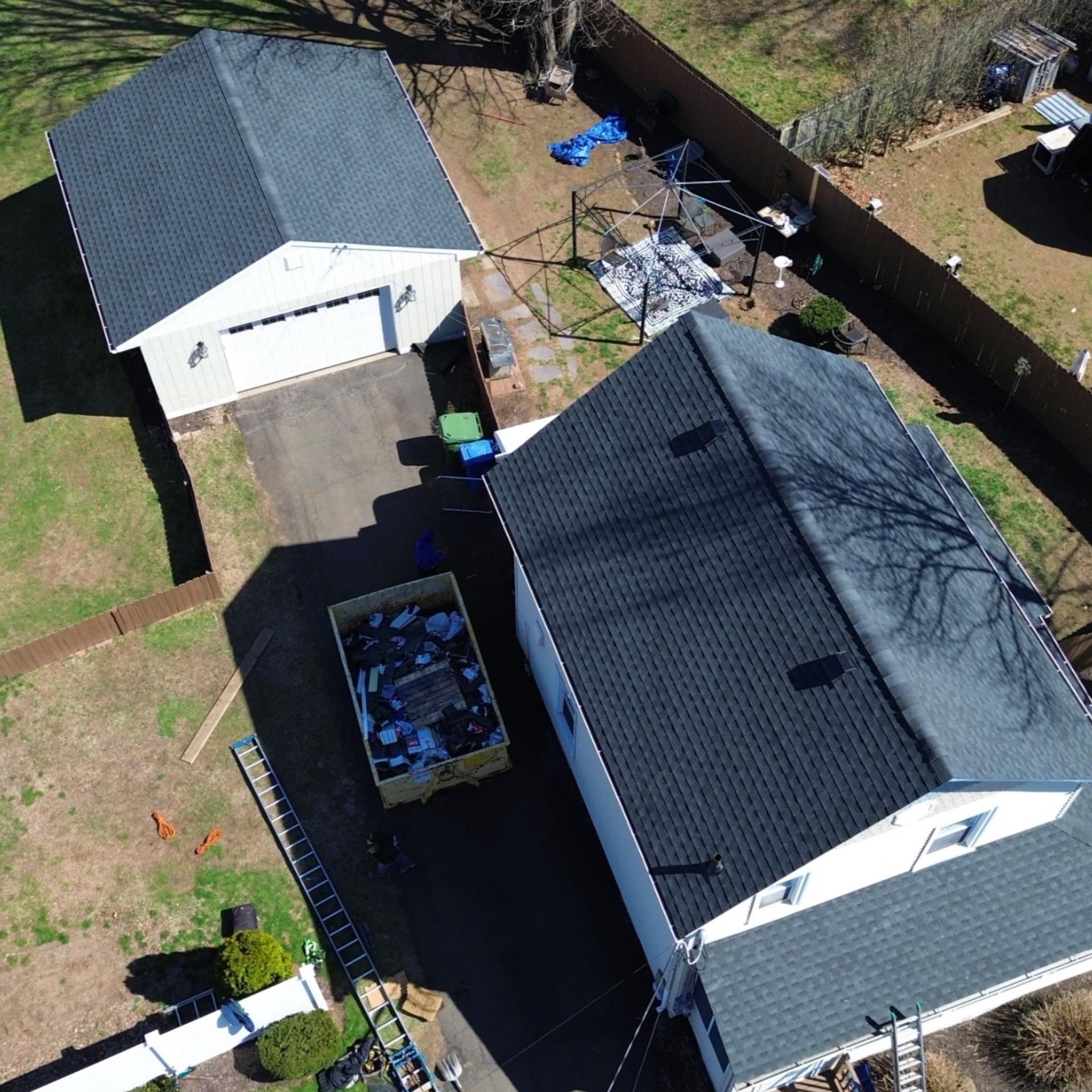 Aerial view of two white houses with dark roofs, a driveway, and a backyard with scattered debris.