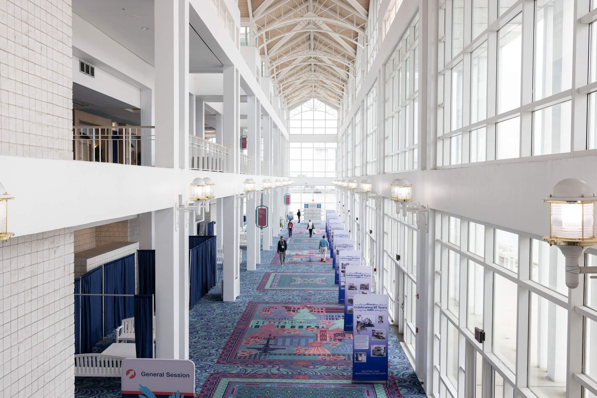Bright convention center corridor with colorful carpet, tall windows, and a high arched ceiling; a few people walk through.