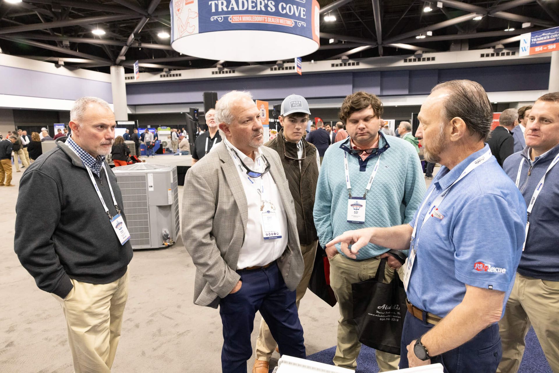 Four men talking at a trade show booth, one in a blue polo holding a brochure.