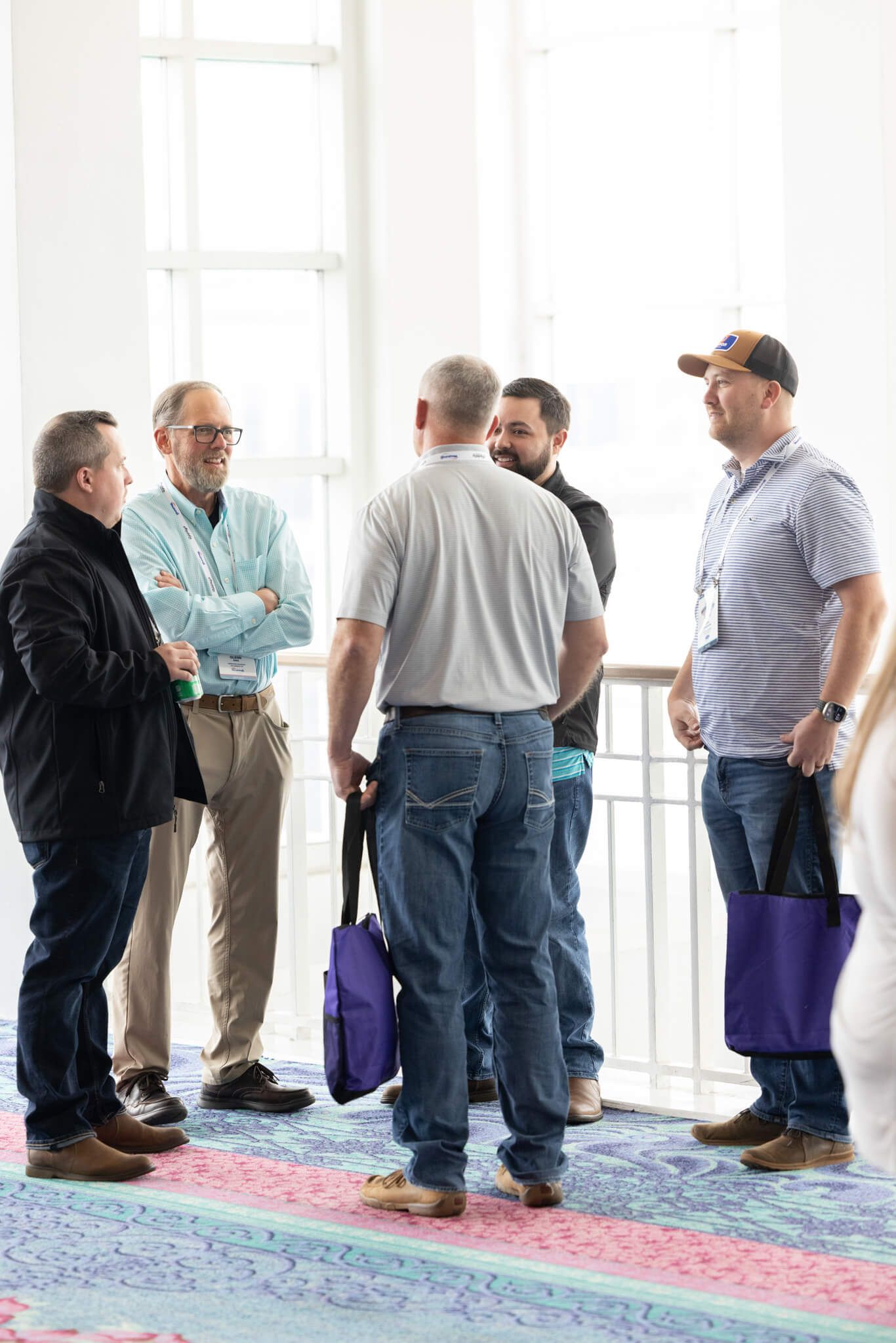 Five men stand chatting in a bright room near tall windows, with purple bags on the floor.