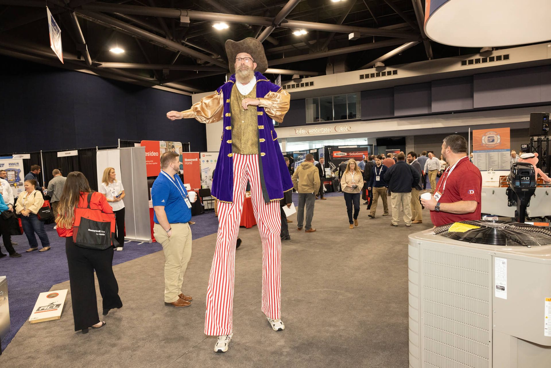 Tall stilt performer in a purple jacket and red-striped pants at an indoor event hall, surrounded by attendees.