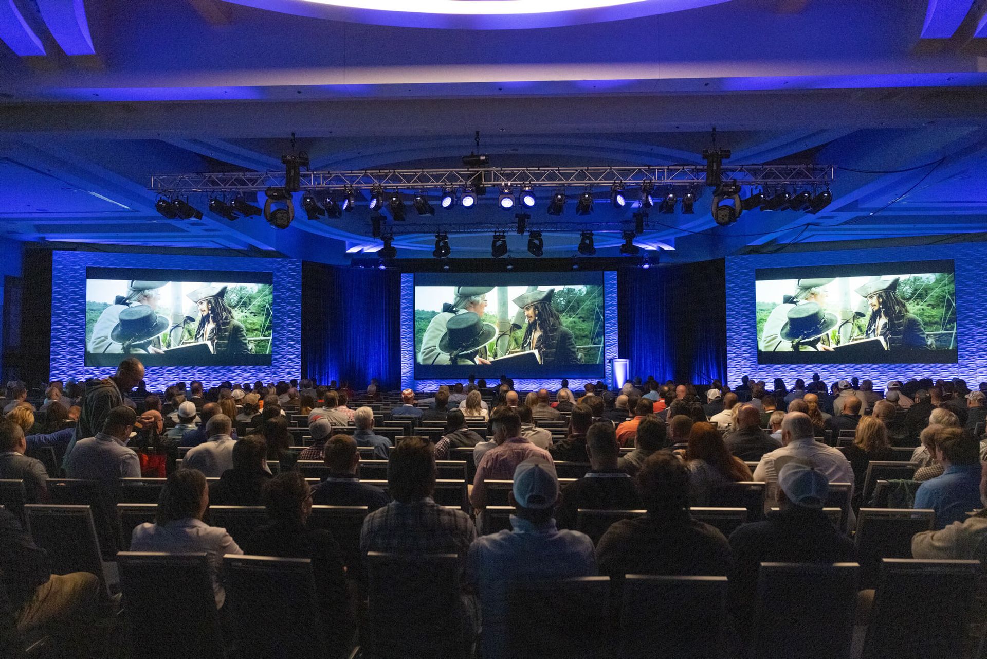 Audience at a blue-lit conference watching a presentation on large screens in a ballroom