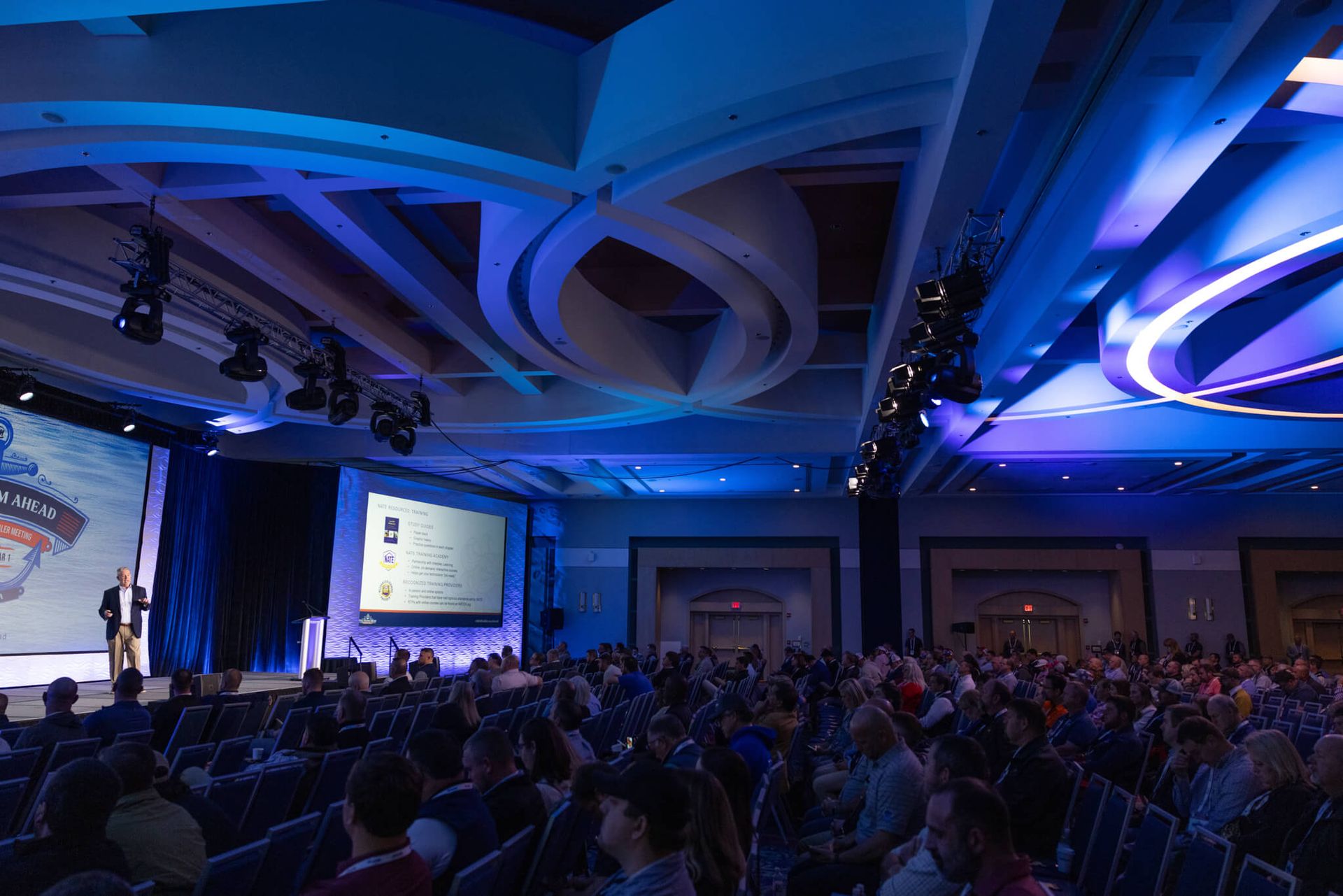 Conference hall with blue lighting, a seated audience, and a presentation screen on stage