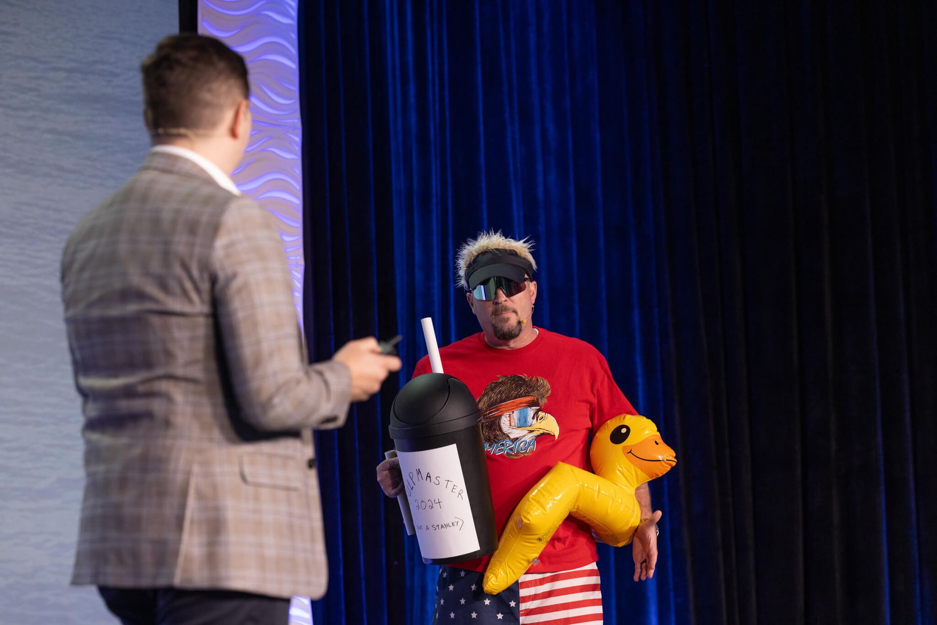 Man on stage in plaid jacket facing another man holding a yellow inflatable toy and white bucket against blue curtains