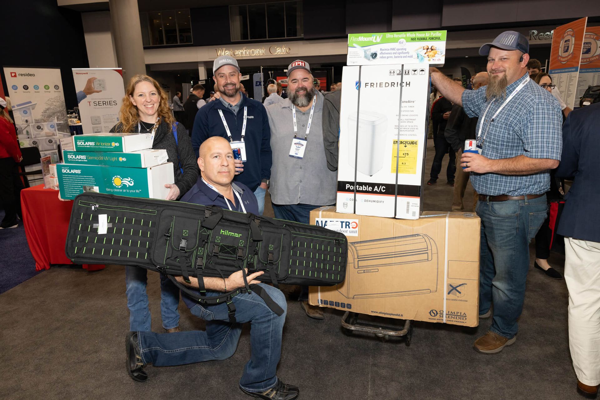 Group posing at an indoor event with boxed products and a large black case on a rolling cart.
