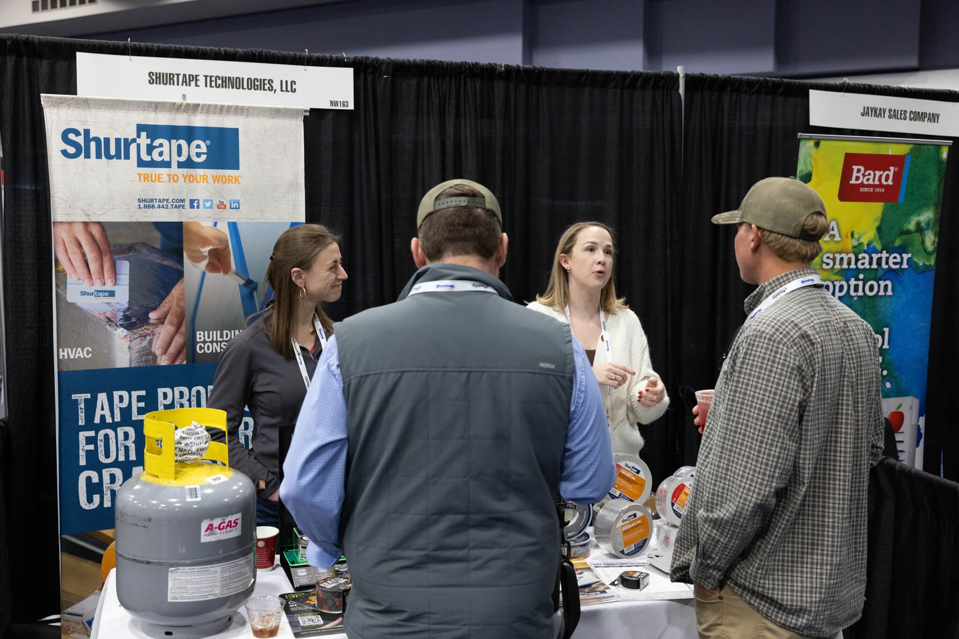 Booth staff talking with attendees at a trade show display for Shurtape and Susi brands.