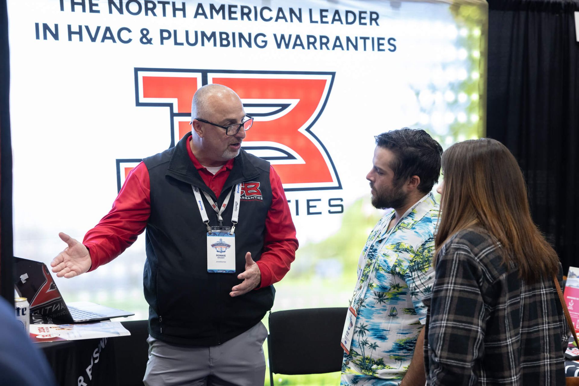 Three people talking at a trade show booth with a red logo and HVAC plumbing warranty banner