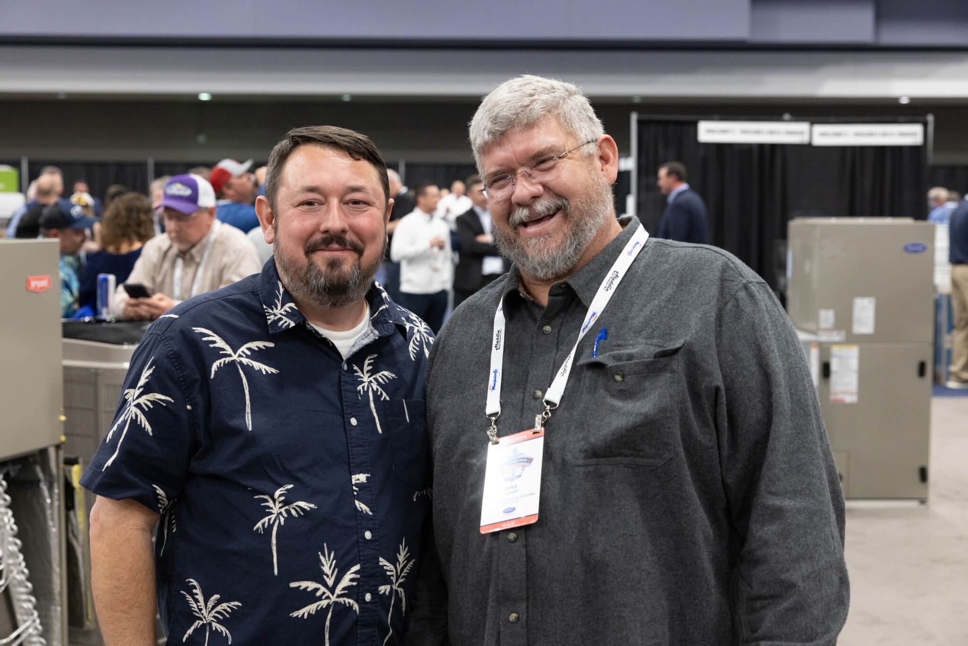 Two men smiling at a conference, wearing lanyards in an expo hall.