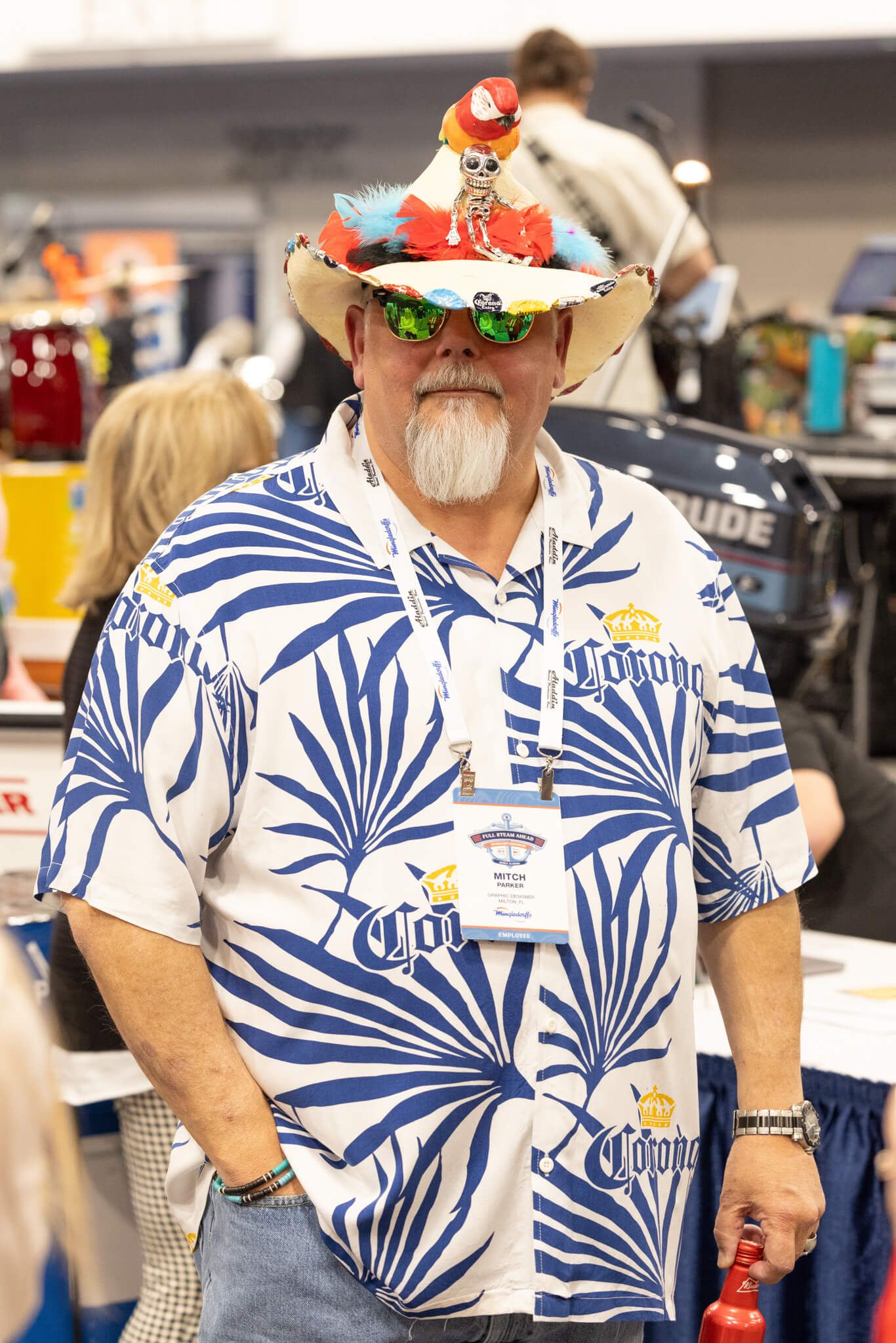 Man in a blue tropical-print shirt wearing a festive hat, standing indoors at an event booth