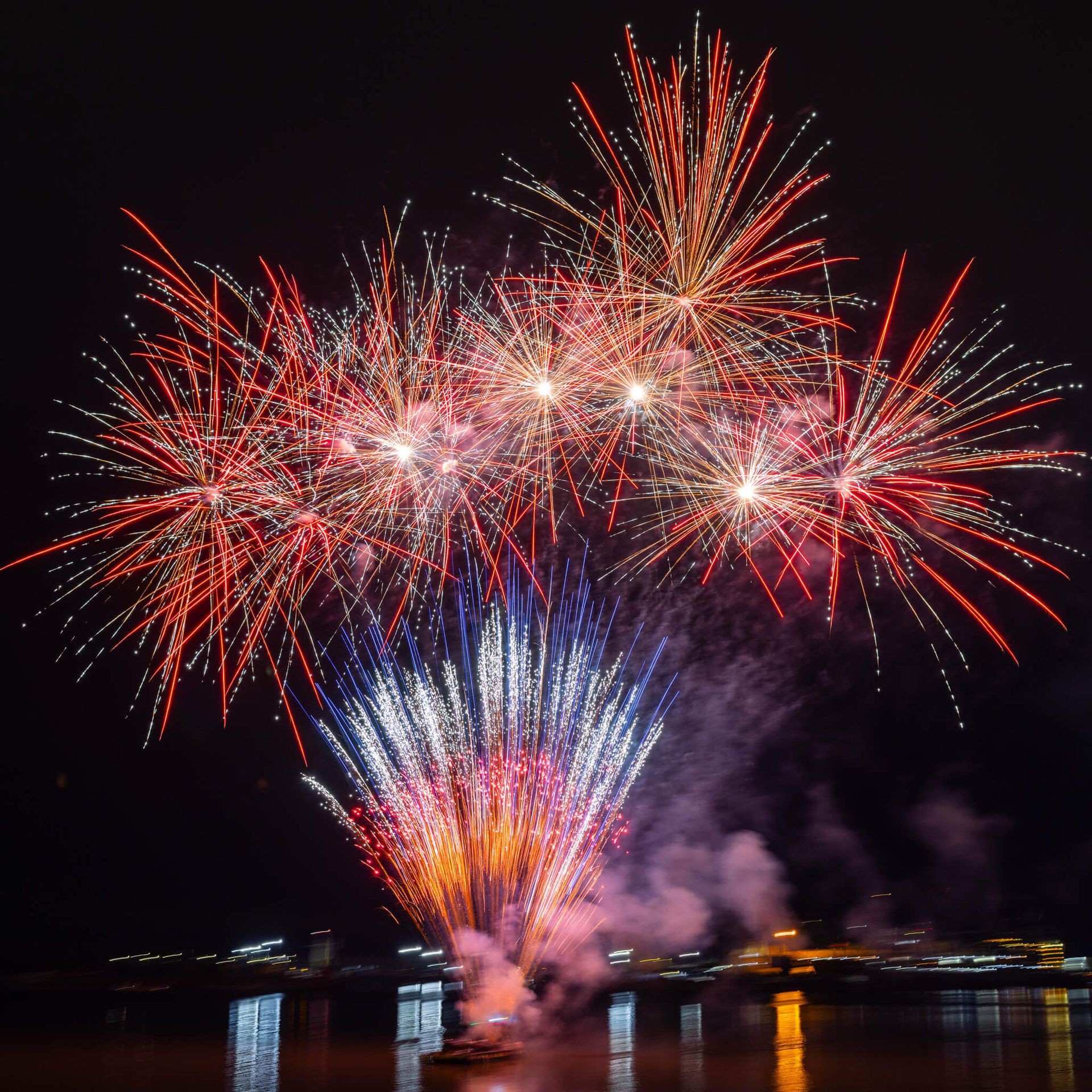 Colorful fireworks bursting over water at night, with red, blue, and gold sparks