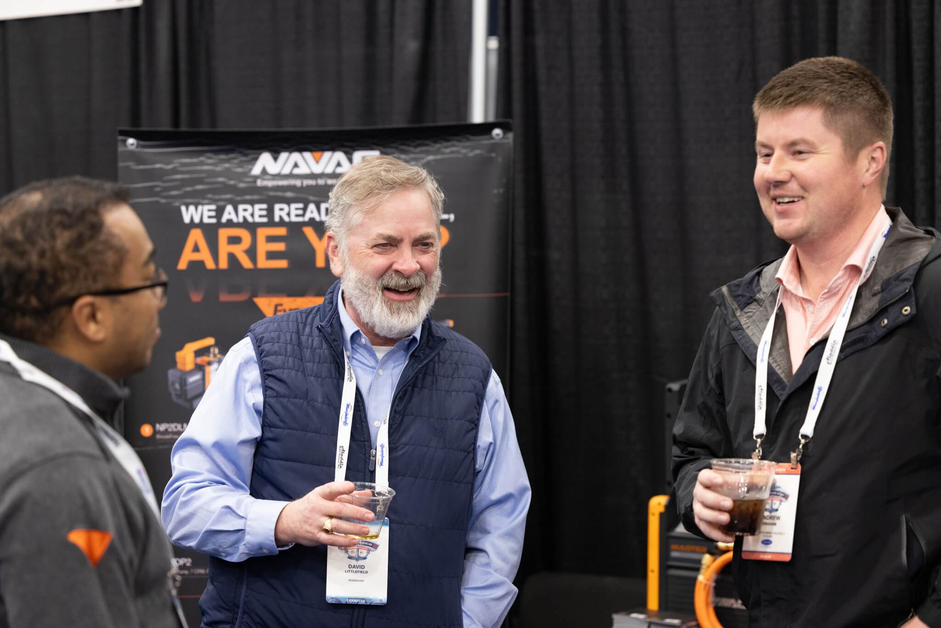 Two men talking at a trade show booth, holding drinks and smiling in front of a black banner.
