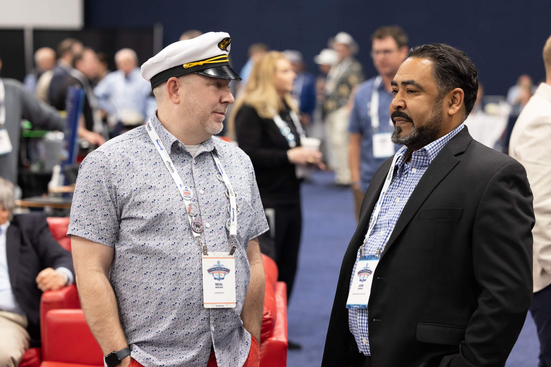 Two men in conversation at a conference, wearing lanyards in a crowded event hall.