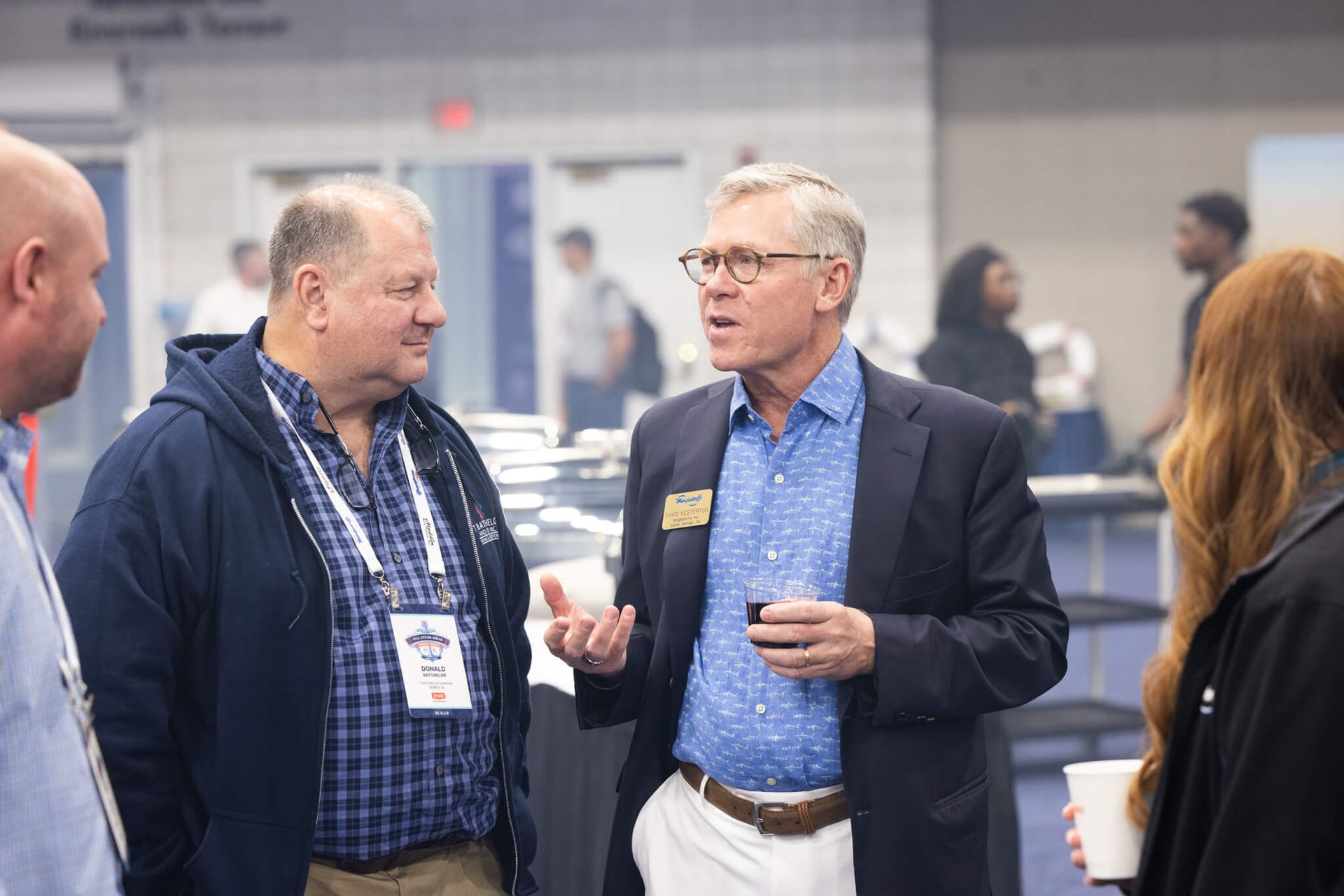 Two men and a woman talking at a conference booth, with one man holding a drink and wearing a name badge.
