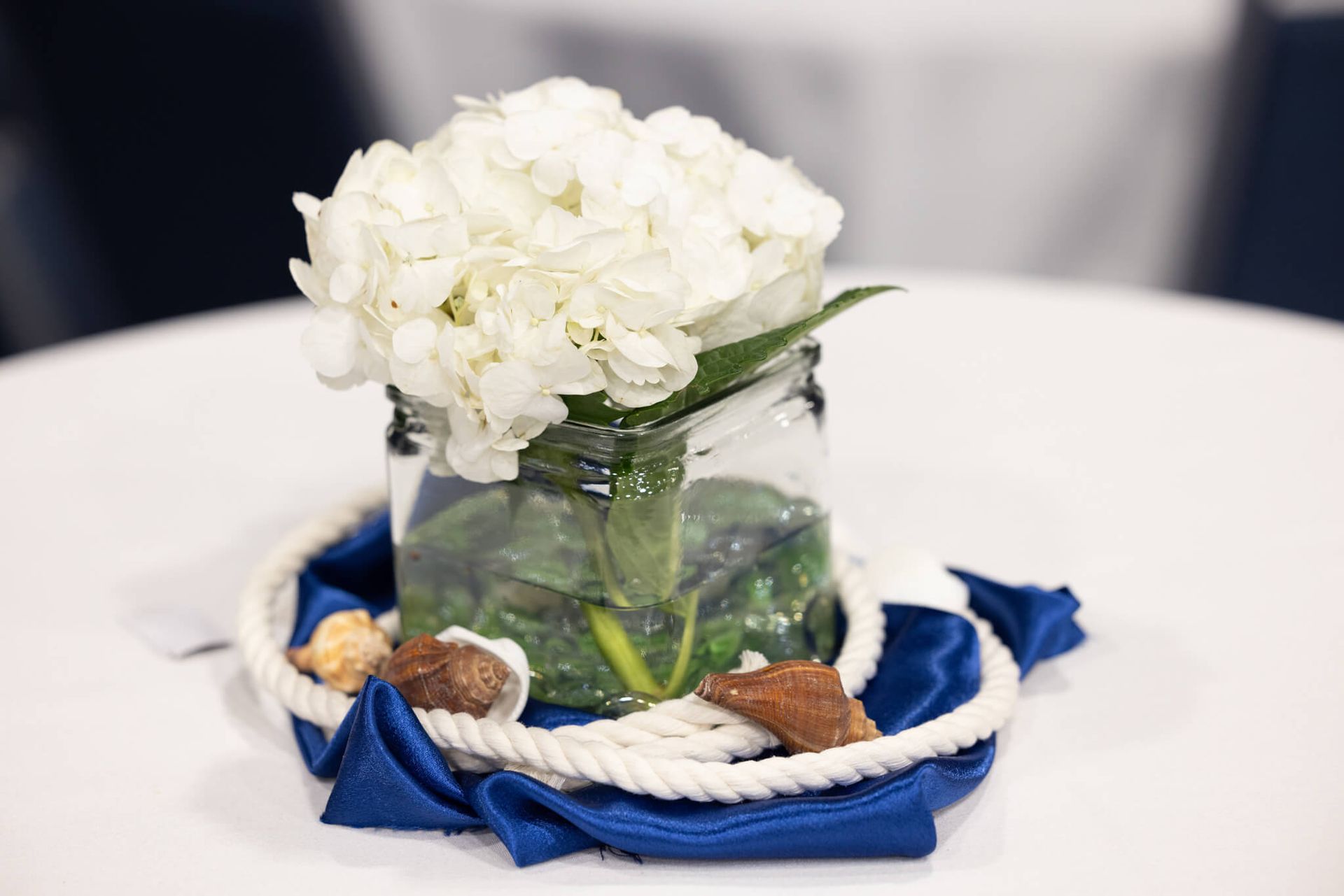 White flowers in a glass jar on a blue napkin centerpiece with seashells on a table.
