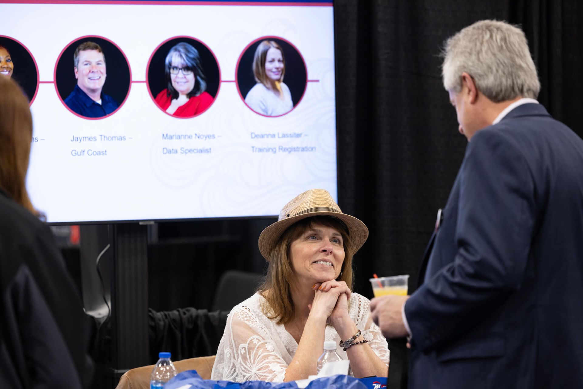 Woman in a hat listening at a conference table while a presentation screen shows speaker headshots.