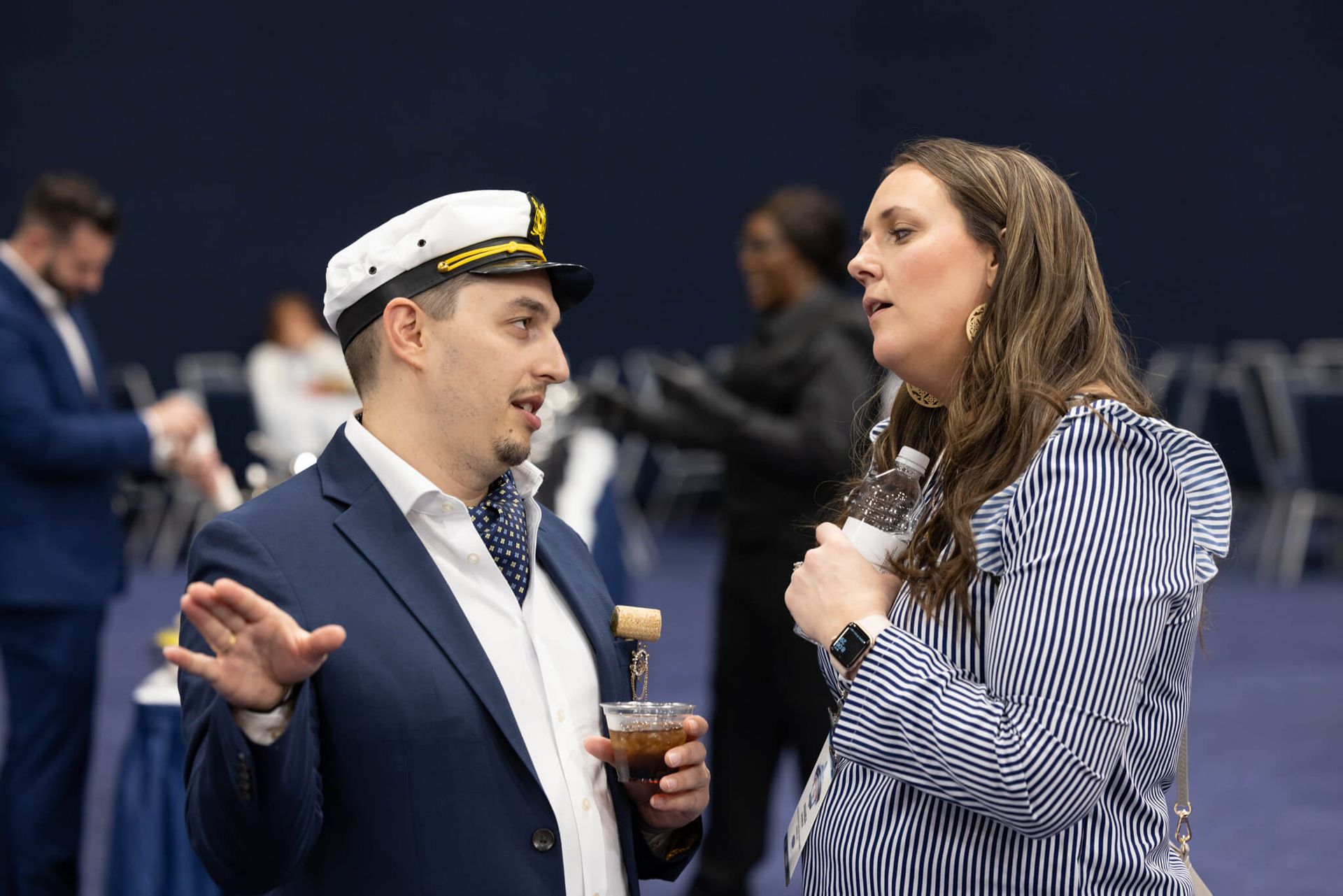 Two people chatting at an event, one in a captain’s hat holding a drink and the other in a striped blazer.