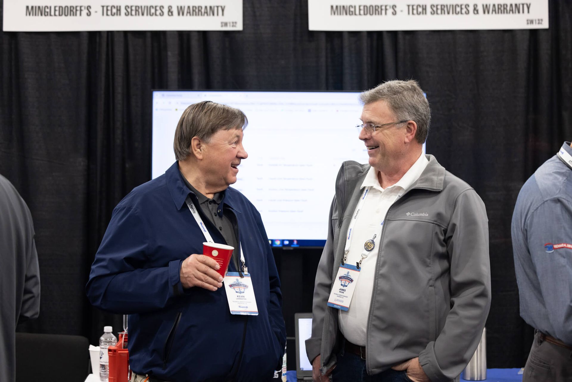 Two men talking at a trade show booth with banners and a monitor behind them
