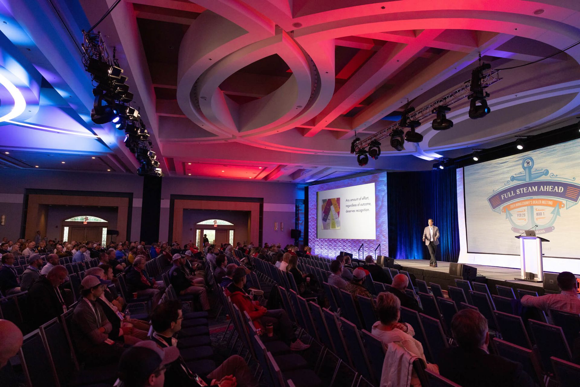 Audience seated in a ballroom conference hall with red and blue stage lighting and a large presentation screen