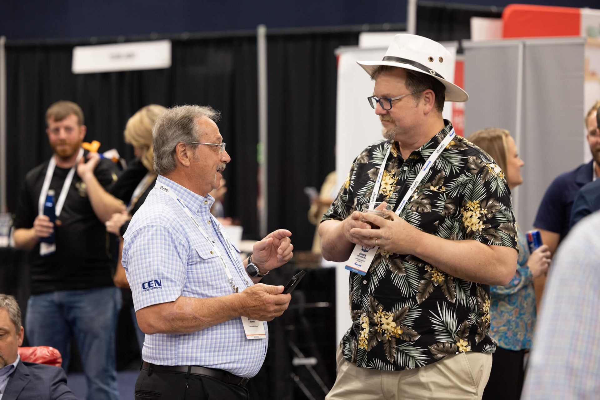Two men talking at a conference, one in a white hat and floral shirt, with attendees and booths in the background.