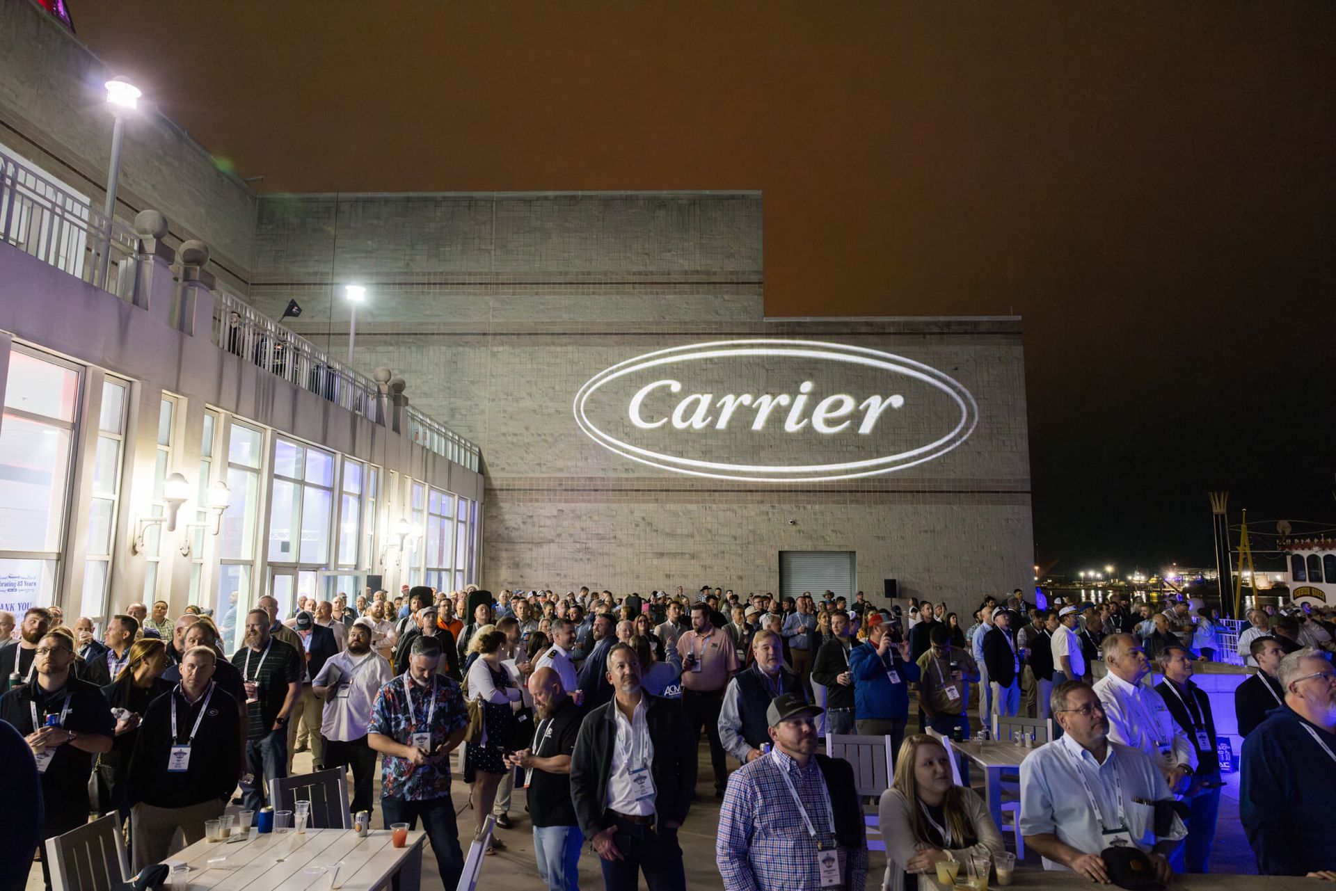 Crowd gathered outside a Carrier building at night, with bright lights and a large Carrier logo on the wall.