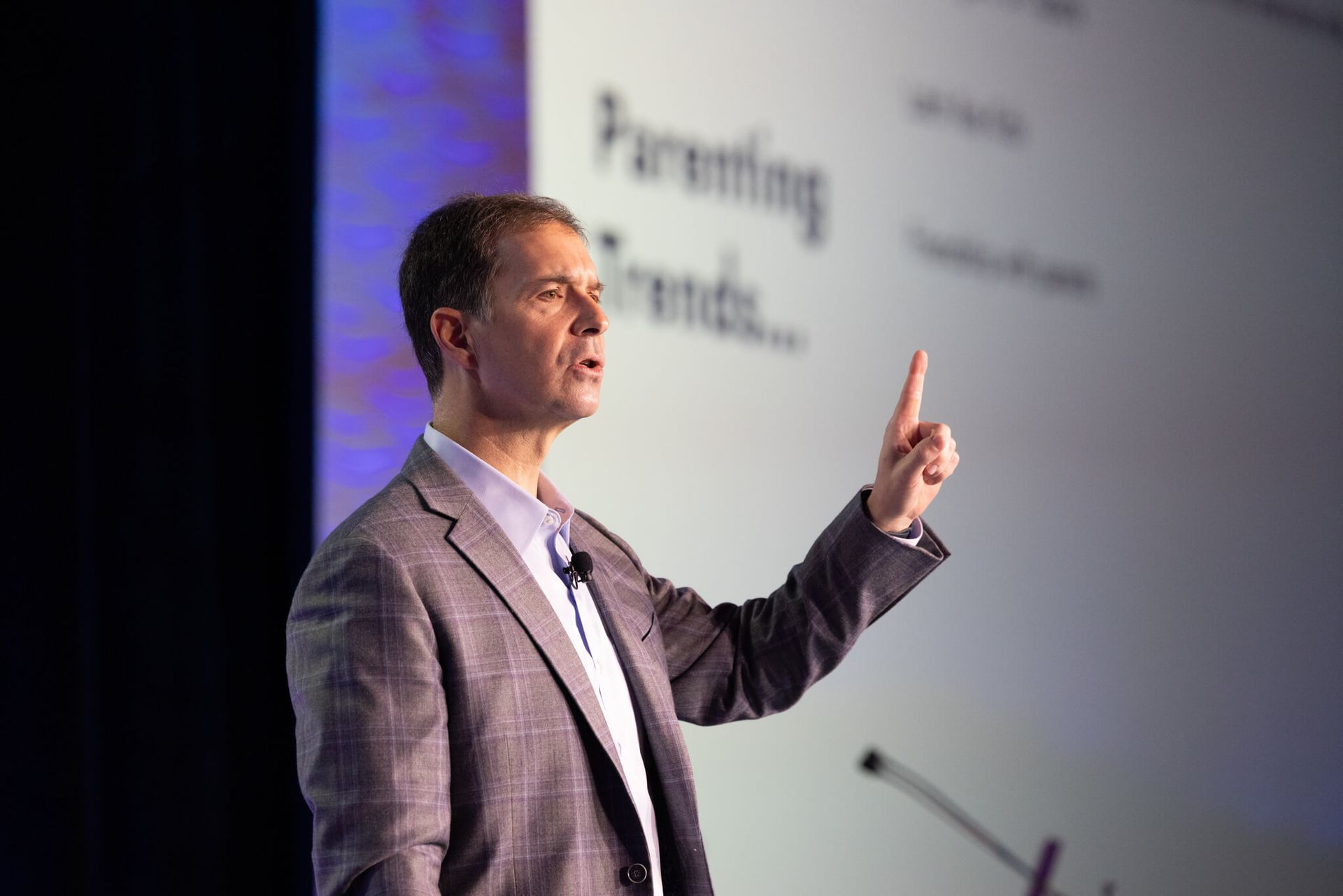 Presenter in a gray suit speaking onstage and pointing at a projected screen