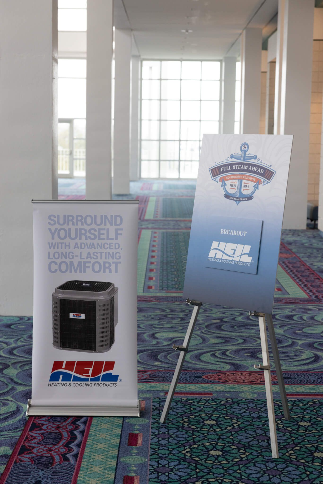 Two promotional signs on stands in a hallway, one gray and one white, with a carpeted floor and bright windows.