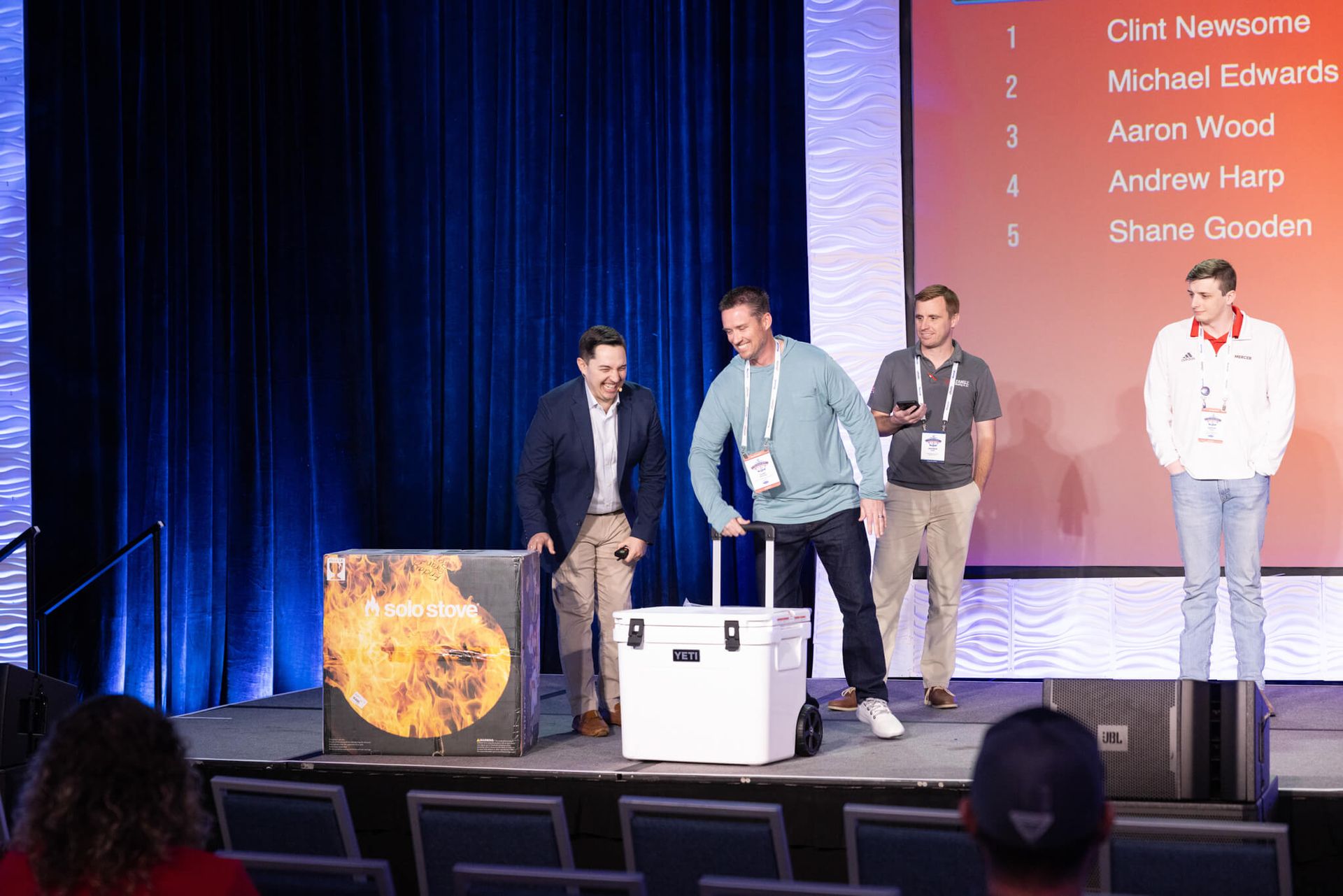 Four men onstage at an awards event, standing by a podium with a screen listing names behind them.